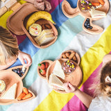 Photo prise ne hauteur d'un pique nique d'enfants. les enfants sont assis sur un plaid coloré avec plusieurs assiettes et planches en bois posé dessus avec différents aliments.