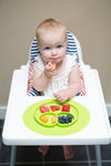 Photo d'un bébé assit sur sa chaise haute, entrain de manger un bout de carotte avec ses doigts. Sur le plateau de sa chaise haute est posé un plateau avec assiette compartimentée intégrée. Son assiette est de couleur verte