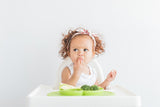 Photo d'une petite fille rousse au cheveuc bouclées qui est assise sur une chaise haute et qui mange avec ses doigts. Devant elle est posée son assiette de couleur verte