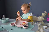 Jeune fille assise à table, elle est souriante et joue avec une marionnette en bois. Devant elle sur la table est posée de la vaisselle en silicone, dont une assiette compartimentée avec des fruits rouges dedans.
