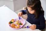 Photo d'une jeune fille qui mange à table. Elle a des couverts de couleur violet, et elle coupe des aliments disposés dans une assiette de couleur rose. il y a des legumes et des pommes de terre