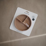 Emballage d'une assiette en silicone compartimentée de la marque Mushie. L'emballage laisse apparaitre l'assiette qui est de couleur beige