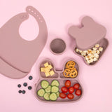 Photo prise en hauteur d'une table rose avec de la vaisselle pour enfant en silicone de la même couleur. il y a une assiette en formede lapin à 3 compartiments avec des ingrédients dedans, un gobelet, un bavoir et une boite à gouter avec son couvercle à moitié fermé qui contient des pop corn