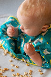 Bébé entrain de mangé des céréales eparpillés devant lui sur un plateau repas. Le bébé est blond et il porte un bavoir de couleur et jaune avec des illustrations de zèbres