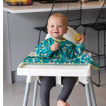 Bébé assit sur une chaise haute, il est souriant et regarde l'objectif, il tient dans sa main un epis de maïs. Il est habillé d'un bavoir intégral à manche longue de couleur vert et jaune avec des illustration de zèbres