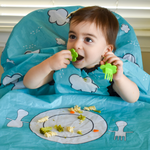 Photo d'un enfant entrain de manger, il est assit sur une chaise haute qui est recouvert d'un tablier de couleu vert avec des illustration blanche en forme de nuage. A l'emplacement de la tablette il y a une illusatrtion de couverts et d'assiette ou sont posés directement les aliments dessus: pate et carotte