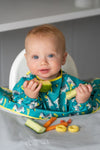 Bébé entrain de mangé des légumes eparpillés devant lui sur un plateau repas. Le bébé est blond et il porte un bavoir de couleur et jaune avec des illustrations de zèbres
