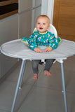 Bébé assit sur une chaise haute équipé d'un maxi plateau en tissu de couleur gris. Le bébé a un tablier a manches longues accroché au plateau. Son bavoir est de couleur bleu avec des coutures jaunes et des illustrations de zèbres