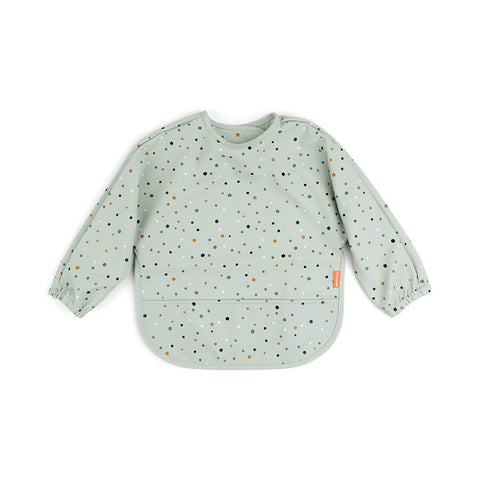 Sur fond blanc, un tablier pour enfant aux manches longues avec une poche centrale. Le tablier est vert clair avec des petits ronds comme des confettis aux couleurs beige et marron