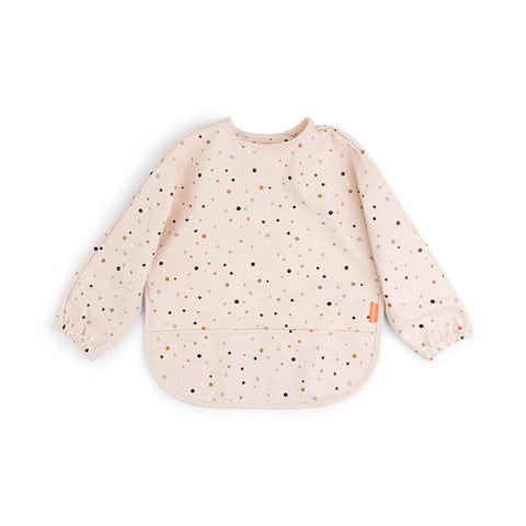Sur fond blanc, un tablier pour enfant aux manches longues avec une poche centrale. Le tablier est rose pâle avec des petits ronds comme des confettis aux couleurs beige et marron