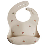Photo sur fond blanc d'un bavoir en silicone de couleur beige avec des motifs arc en ciel. Le bavoir est ajustable et il a une poche de recupération sur le devant