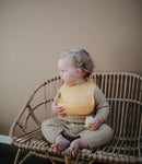 Jeune enfant assi sur un banc en rotin. Il est habillé d'une combinaison beige et il porte autour de son coup un bavoir en silicone de couleur orange avec des motifs blanc