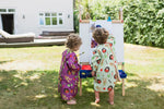Photo de 2 jeunes enfants qui font de la peinture sur un chevalet dans un jardin. Les enfant ont tous les 2 des tabliers assez long de couleurs differente, l'un est violet et l'autre vert pistache.