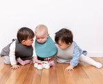 Photo de 3 bébés assient par terre, ils portent chacun un bavoir en silicone de couleur gris, vert et gris clair. Ils sont assis les un a coté des autres et devant le 2emem bébé est posé une assiette en forme de lapin avec des morceaux de fraises dedans