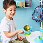 jeune garcons qui souris et tient dans sa main droite une boite à repas avec des poignées bleus, sa boite contient des pates et des légumes