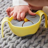 Gros plan de la main d'un bébé entrain de prendre des gâteaux dans un bol de couleur jaune avec un couvercle anti chute de couleur gris