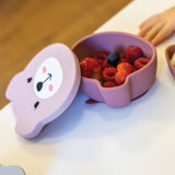 Photo d’une table à manger avec posé dessus de la vaisselle pour enfant en silicone de couleur rose. En avant plan il y a un Bol pour enfant de couleur rose avec une forme de tête d’ours. l est équipé d’ couvercle de couleur rose clair avec des illustrations représentant le visage d’un ours. Dans le bol il y a des fruits rouges