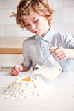 Petit garçon qui verse du lait a l'aide d'un bol transparant sur un tas de farine formé sur un plan de travaille de cuisine. Le tas de farine forme un puit avec un oeuf à l'intérieur
