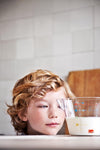 Photo d'un petit garçon ou l'on voit juste sa tête posée sur une table qui regarde un bol mesureur avec du lait à l'intérieur