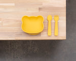 Photo d'un bout de table en bois avec posé dessus une bol de couleur jaune en forme de tête d'ours et à coté une cuillère ainsi qu'une fourchette assortis. Ce sont des vaisselles pour enfant