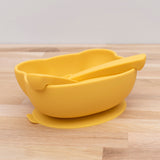 Photo d'un bol avec ventouse de couleur jaune posé sur une table en bois, à l'interieur est posé deux couverts avec des embouts arrondis. Les couverts sont de la même couleur que le bol et ils sont en silicone comme le bol