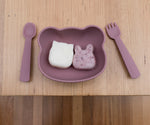 Photo d'une table en bois avec posé dessus une assiette pour enfant en forme de tête d'ours avec a l'intérieur 2 gouter en forme d'animaux, a coté du bol sont posés une cuillère et une fourchette assortient à l'assiette