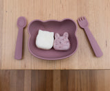 Photo d'une table en bois avec posé dessus une assiette pour enfant en forme de tête d'ours avec a l'intérieur 2 gouter en forme d'animaux, a coté du bol sont posés une cuillère et une fourchette assortient à l'assiette