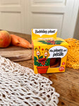 Photo d'un emballage en carton de couleur jaune d'une plante à planter. La plante s'appelle ma plante qui pétille. Elle est posée sur une table en bois avec des carottes en arrière plan