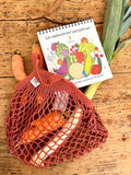Filet de course de couleur rouge, contenant des carottes et un poireau. il est posé sur un plan de travail en bois. un calendrier perpétuel illustré de fruit et légumes rigolos sort également du sac à provisions