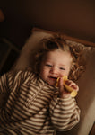 Photo d'un jeune enfant allongé sur une table à langer, il tient dans sa bouche un jouet de dentition en forme de brosse à dents. La photo a des ton ocre et beige