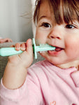 Bébé qui tient dans sa bouche une brosse à dent à double embout de couleur vert menthe. L'enfant est brun et porte un bodi de couleur rose