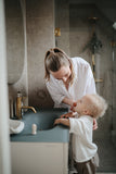 Photo d'une maman et son enfants, dans une salle d bain. la maman est enttrain de brosser les dents du jeune garçon avec une brosse à doigt 