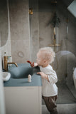 Jeune garçon dans une salle de bain, il tient sur son doigt une brosse à dents à doits. Le garçon a les cheveux blond et frisés