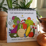 Calendrier perpétuel de fruits et légumes posé sur un chevalet avec des spirales noires. Il est posé sur une table de cuisine, sa 1ere page représente des fruits et légumes illustré de façon rigolote avec des visages