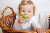 Photo d'un bébé blond au yeux bleu habillé d'un body blanc. Le bébé est dans un panier en osier et il tient dans sa bouche un jouet de dentition en forme de trèfle à 4 feuilles