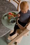 Photo prise en hauteur d'un enfant assit à table, devant lui est posé un tapis en silicone avec de la vaisselle de la même couleur verte olive