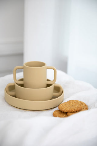 Ensemble de vaisselle pour enfant en silicone de couleur beige, osé sur une nape blanche avec des cookies posés à coté. La vaisselle est empilée les un dans les autres, il y a un assiette, un bol et une tasse