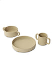 Vaisselle en silicone pour enfant sur un fond blanc, il y 3 objets une assiette, un bol et une tasse à anses. La vaisselle est de couleur beige