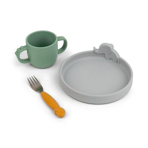 Ensemble de vaisselle en silicone posé sur un fond blanc. il y a une assiette en silicone de couleur grise avec sur un rebord un élephant. Une fourchette avec un manche en silicone de couleur moutarde et une tasse de couleur verte avec 2 ansses