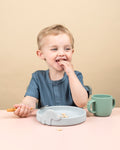 Jeune garçon assi devant une table entrain de porter de la nourriture a s sa bouche. il porte un polo de couleur bleu. il a devant lui une assiette en silicone de couleur grise avec un élephany sur le dessus