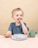 Jeune garçon assi devant une table entrain de porter de la nourriture a s sa bouche. il porte un polo de couleur bleu. il a devant lui une assiette en silicone de couleur grise avec un élephany sur le dessus