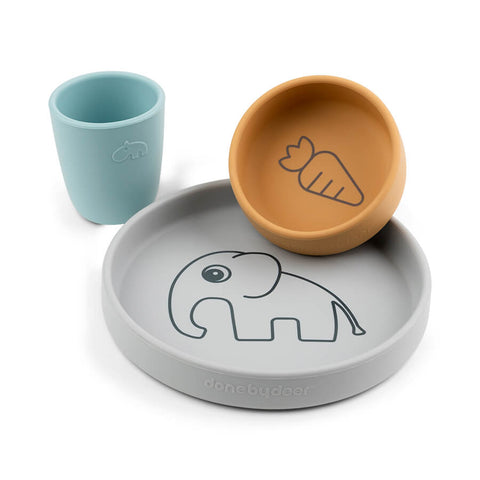 Vaisselles pour enfants sur fond blanc, il y a une assiette de couleur grise avec une illustration d'elephant sur le centre, un bol marron avec une illustration de carotte et un gobelet de couleur bleu