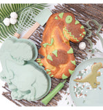 Gâteau en forme de dinosaure décoré avec un glaçage de coueur vert sur quelques élements du gâteau. Le gâteau est posé sur un set de table formé avec des morceaux de bois, il y a a coté de lui des ustensiles de cuisine aux couleur vert pastel et notamment le moule en silicone qui a permis de faire le gâteau