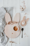 Table de petit déjeuner avec posé dessus 2 planche en bois en forme de tête de lapin, la plus grande contient une tasse de café et à un coeur de gravé à la place du néez. Et la petite est un coquetier contenant un oeuf de couleur blanc, avec des illustration de visage dessus