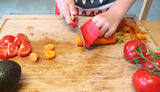 Enfant qui coupe des légumes sur une planche a decouper de cuisine. Il utilise le couteau Petit Chef de la marque Opinel avec son couvre doigt rouge de la marque Opinel. Sur la planche a découper il y a des tomates, une carotte et un avocat