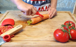 Enfant qui épluches des légumes sur une planche a découper de cuisine. Il utilise l'éplucheur Petit Chef de la marque Opinel avec son couvre doigt rouge de la marque Opinel. Sur la planche a découper il y a des tomates, une carotte et un avocat