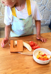 Photo d'un enfant debout devant un plan de travail de cuisine avec posé dessus une planche en bois et des légumes dessus. il y a égalment un coupe legume en bois avec une poignée. L'enfant porte un tablier de couleur bleu clair avec des zébres dessus