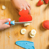 Main d'enfant entrain de couper une banane à l'aide d'un couteau de cuisine pour enfant d'une main et de l'autre un protège doigt rouge en forme de tête d'aimaux avec une toque de chef. sur le bas de la photo il y a un protège lame de couleur bleu posé