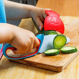 Gros plan sur les mains d'un enfant qui coupe en lammelles un concombre. il tient dans une main un couteau avec une poignée ergonomique et de l'autre un protège doigt de couleur rouge. Il coupe le légume sur une planche en bois 