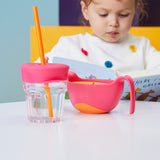 Photo d'une table blanche ou est posé dessus un bol rose avec 2 poignées et un verre avec un couvercle et une paille de la même couleur que le bol. On voit en arrière plan une petite fille qui lit un livre